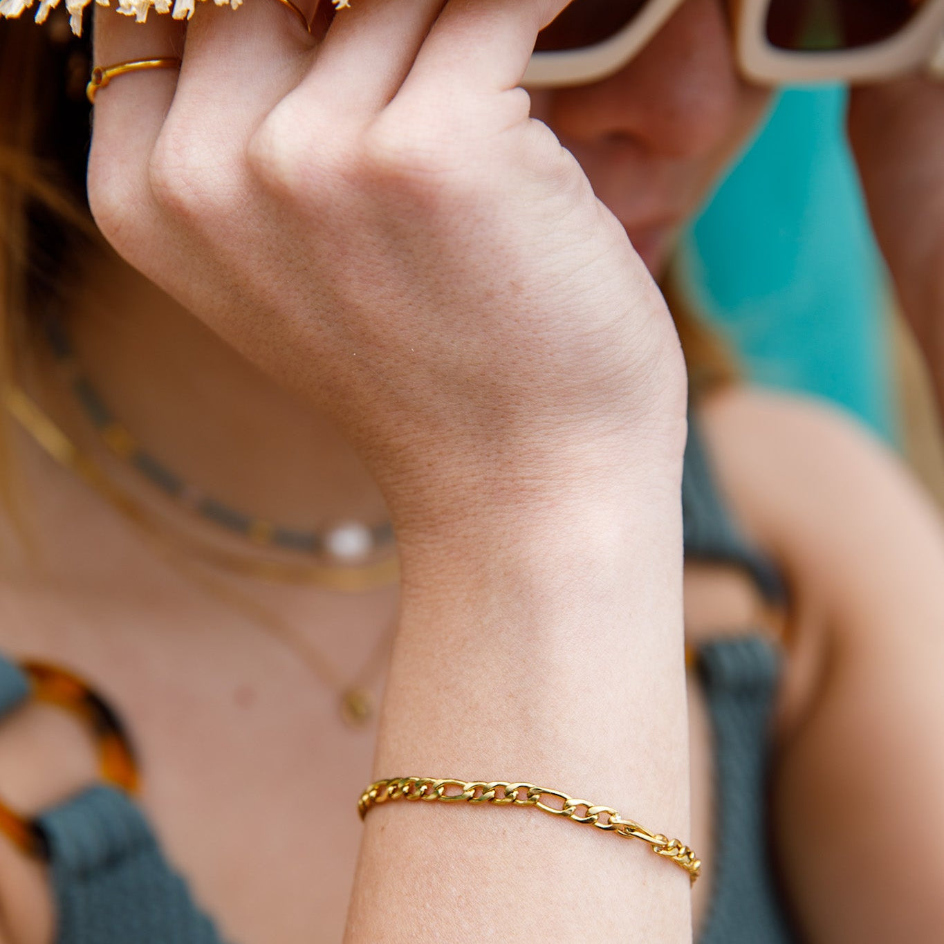 A close-up of a person's wrist wearing a delicate gold chain bracelet, with a straw hat, sunglasses, and a blue top visible in a bright outdoor setting.