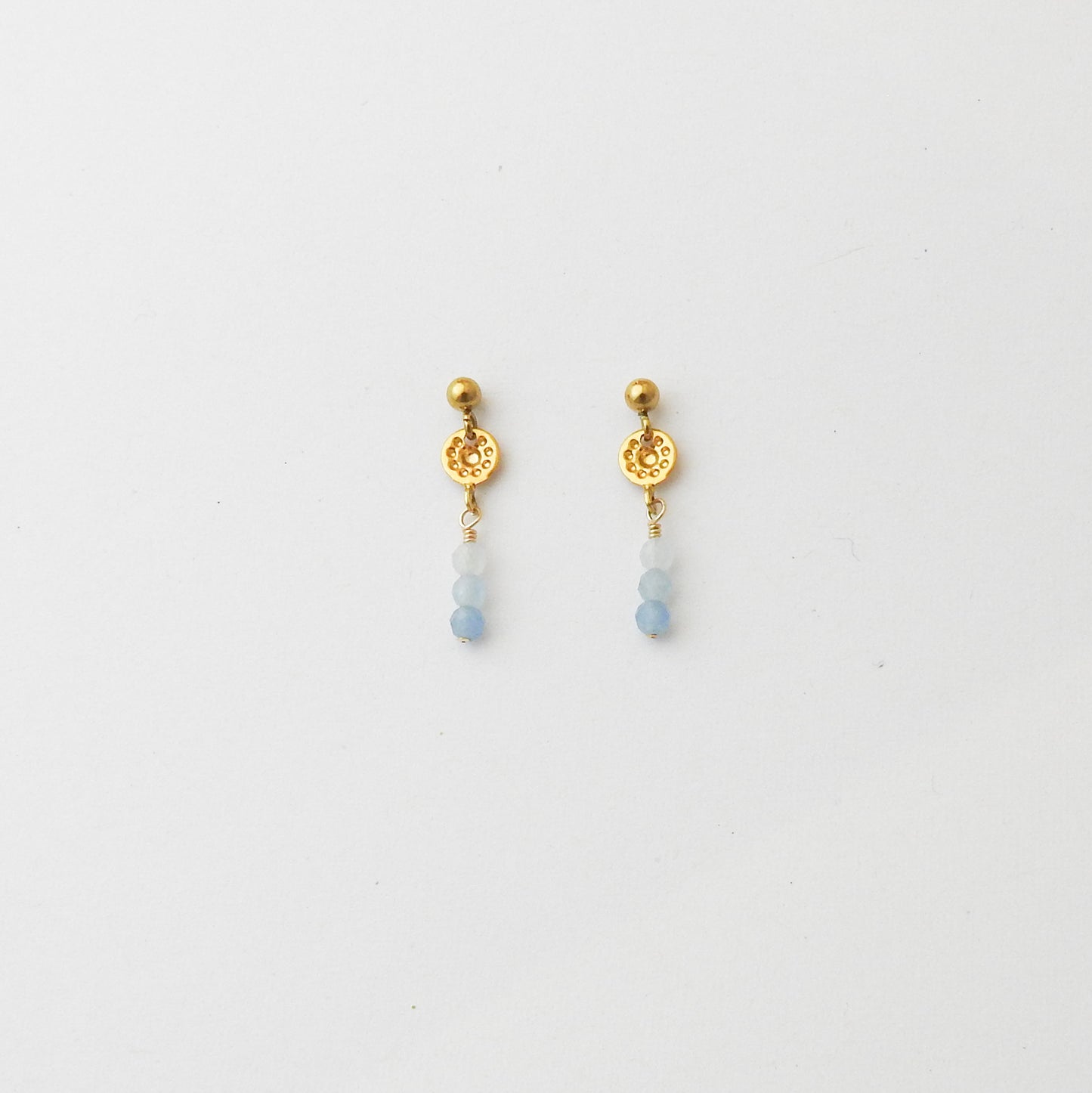 A pair of gold dangle earrings with a textured circular disc, a small gold bead, and three light blue beads hanging below, shown on a plain white background.