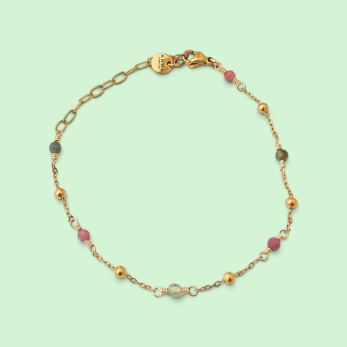 A delicate gold chain bracelet with alternating gold beads, pink gemstone beads, and a small gray-green bead, displayed on a light mint green background.