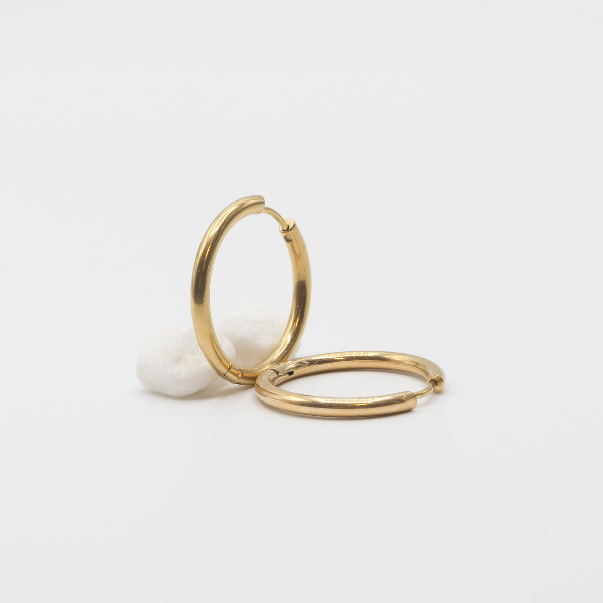 Two gold hoop earrings displayed on a clean white background, with one standing upright and the other lying flat, next to a small white decorative stone.