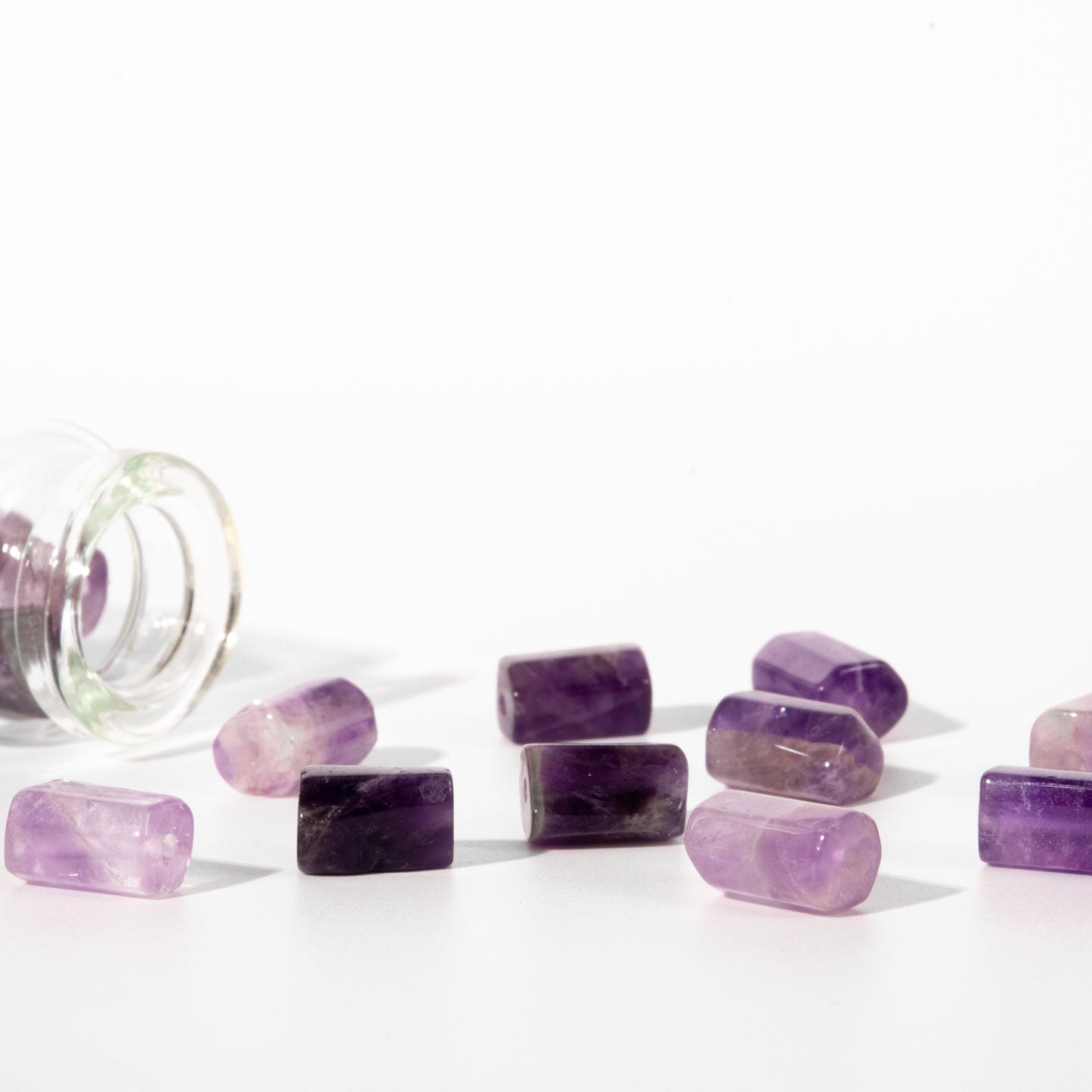 Several polished purple amethyst cuboid gemstones are scattered on a white surface next to an tipped-over clear glass jar, photographed against a bright white background.