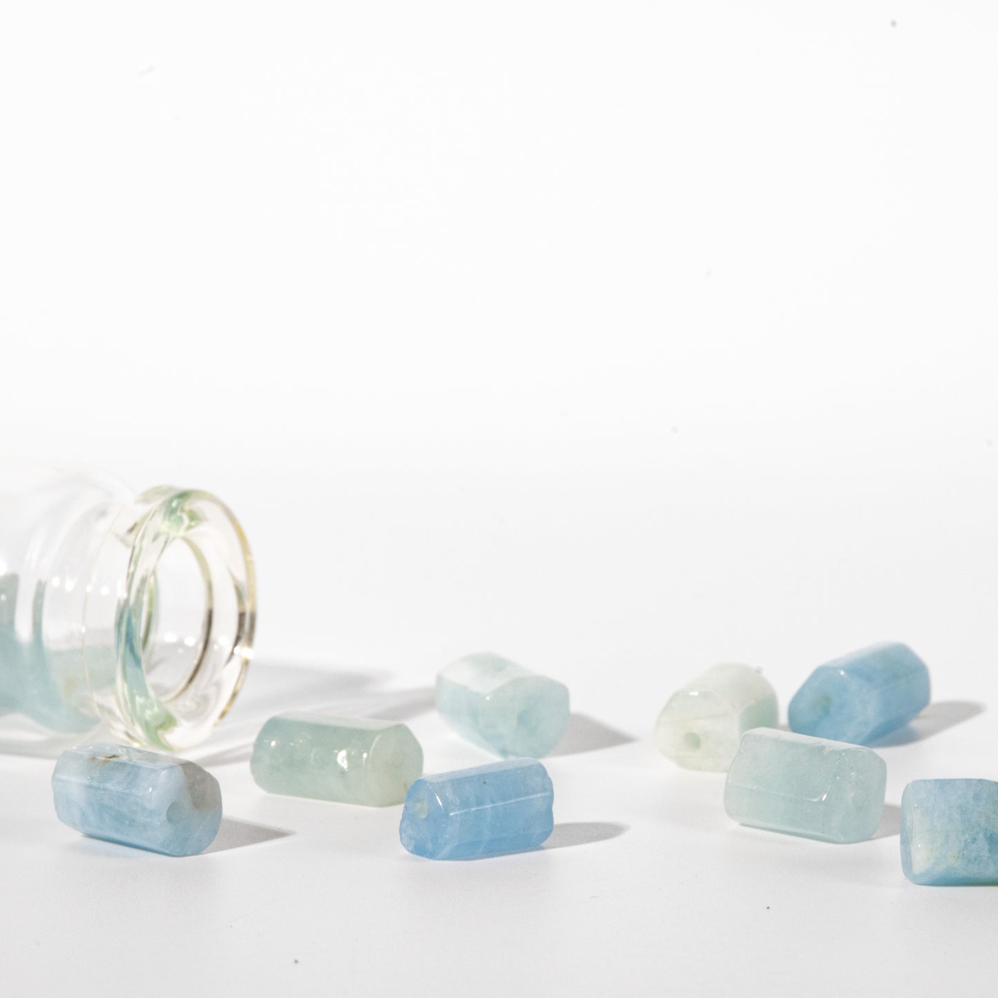 Several small translucent blue and pale green stone beads or charms scattered on a white surface beside a tipped-over clear glass bottle.