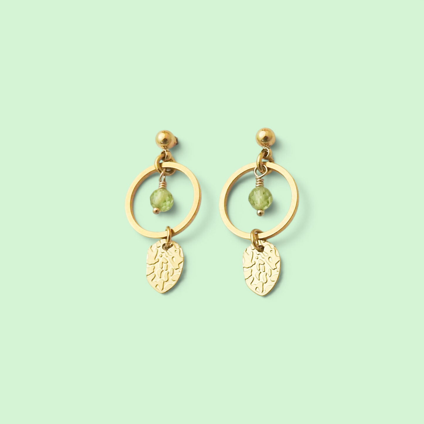 A pair of gold dangle earrings with circular hoops, small bead accents, and leaf-shaped charms, shown on a solid pale mint green background.