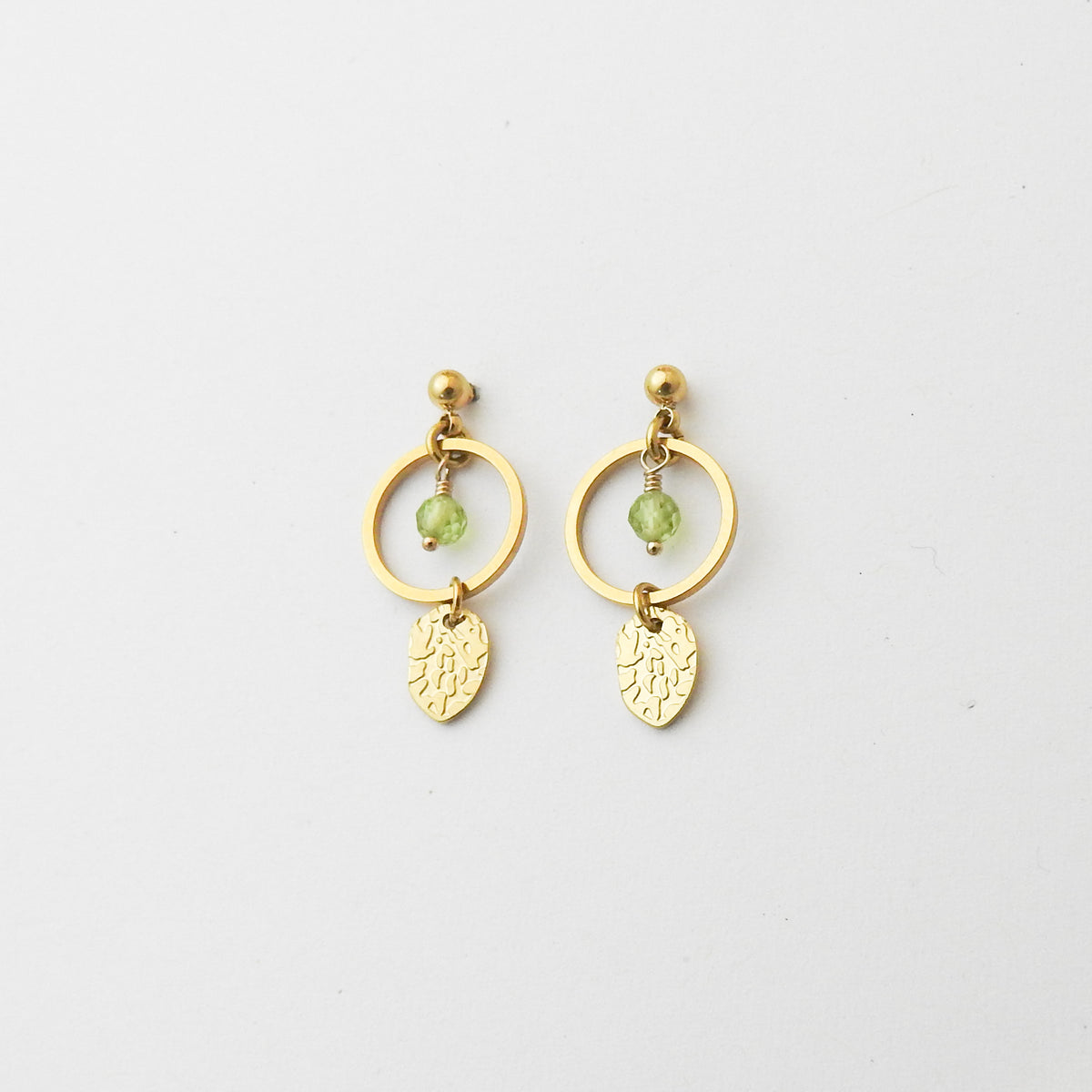 A pair of gold dangle earrings featuring a circular hoop, a small green faceted bead, and a textured leaf-shaped charm, displayed on a clean white background.