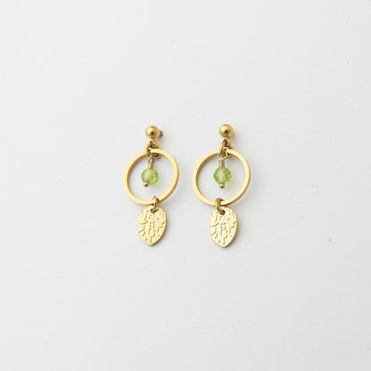 A pair of gold dangle earrings featuring a circular hoop, a small green faceted bead, and a textured leaf-shaped charm, displayed on a clean white background.