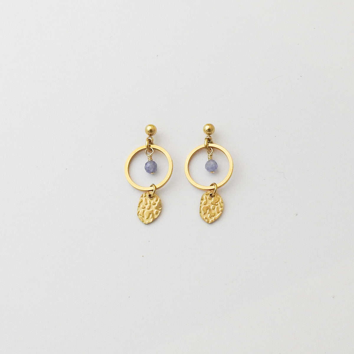 A pair of gold drop earrings featuring a circular ring, a small round purple bead, and a textured gold leaf-shaped dangle, shown on a plain light gray background.