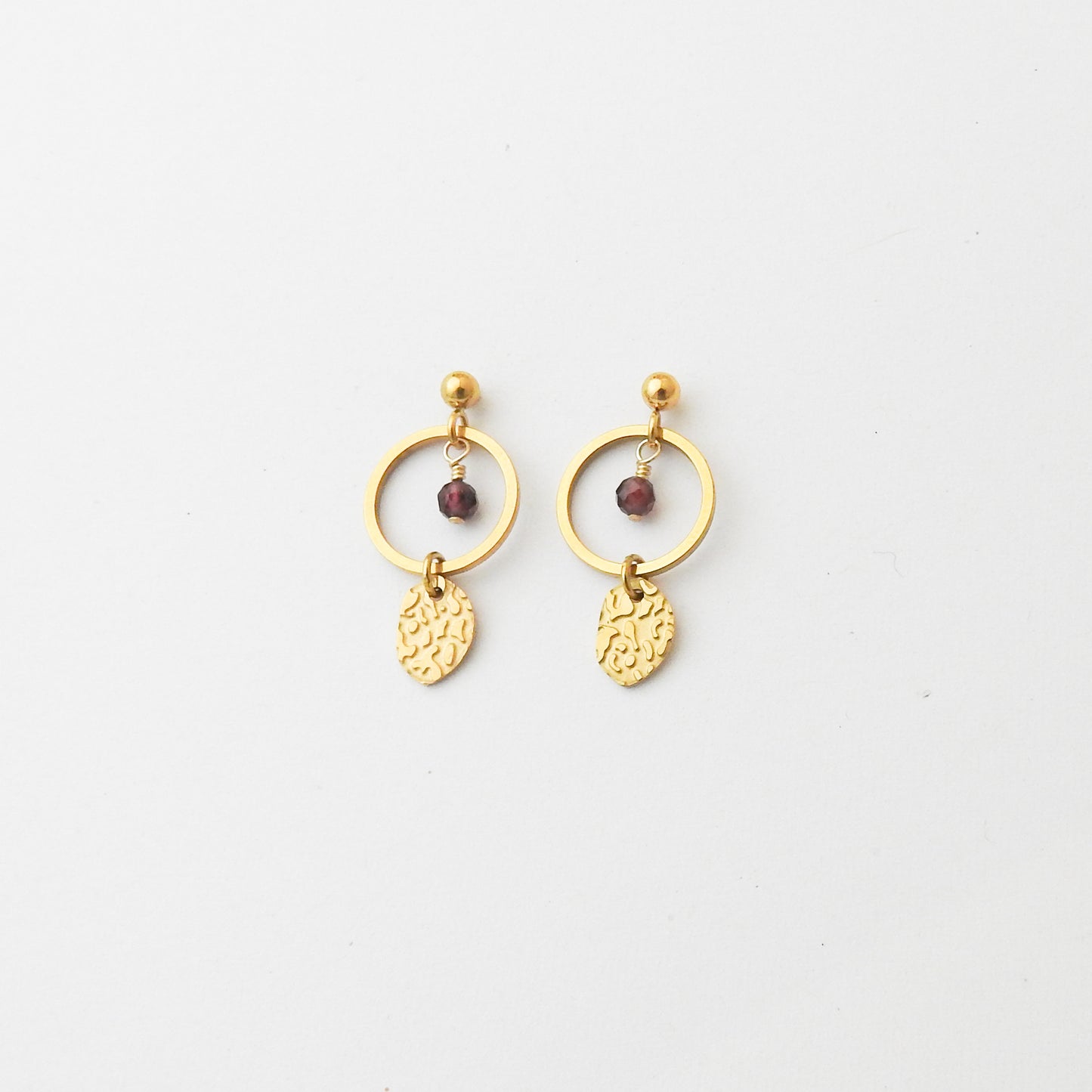 A pair of gold dangle earrings featuring a circular hoop, a small faceted dark red bead, and a textured leaf-shaped charm, displayed on a clean white background.