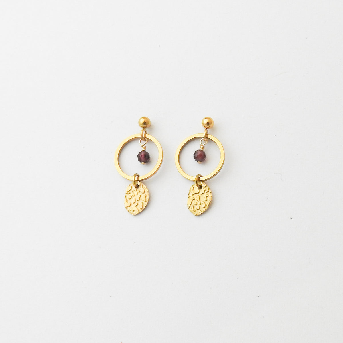 A pair of gold dangle earrings featuring a circular hoop, a small faceted dark red bead, and a textured leaf-shaped charm, displayed on a clean white background.