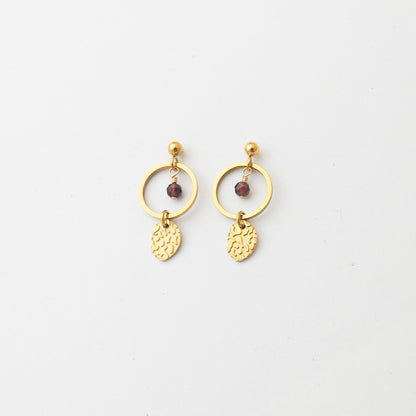 A pair of gold dangle earrings featuring a circular hoop, a small faceted dark red bead, and a textured leaf-shaped charm, displayed on a clean white background.