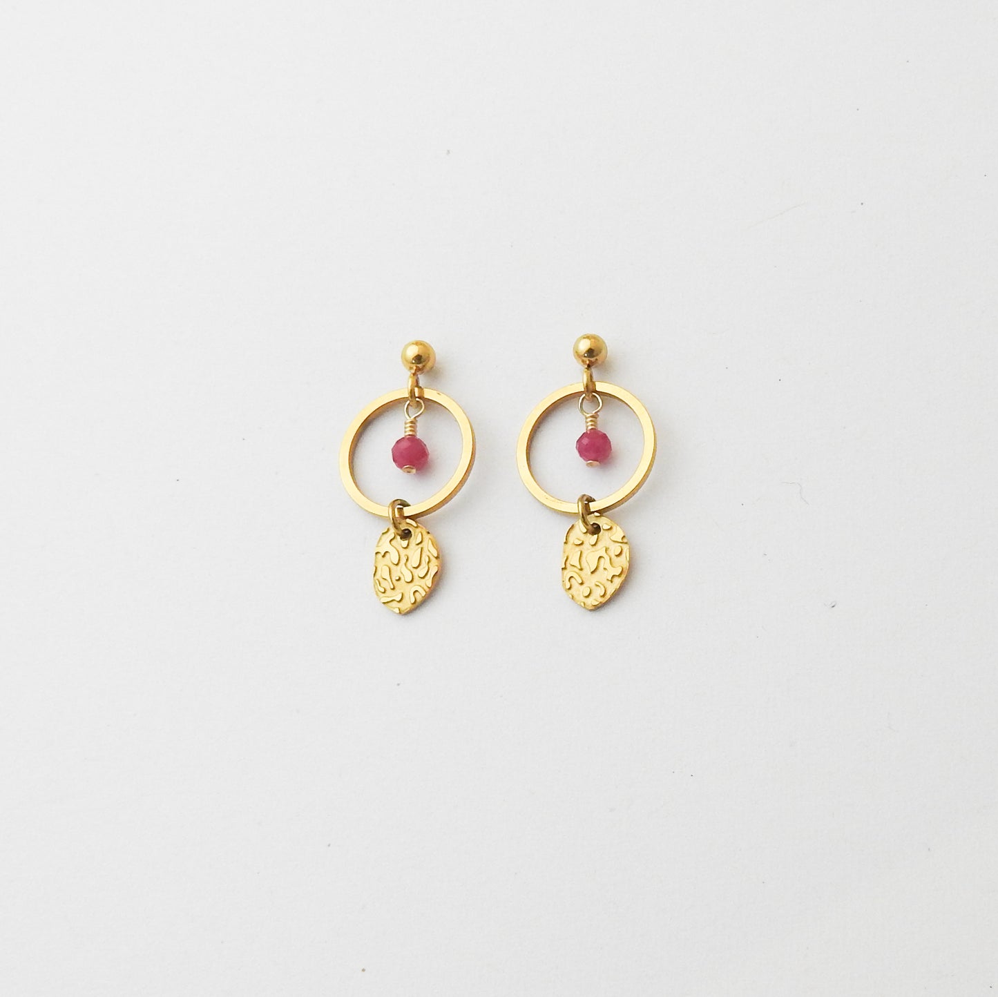 A pair of gold-toned dangle earrings with a circular hoop, a small pink bead, and a textured leaf-shaped charm, displayed on a clean white background.