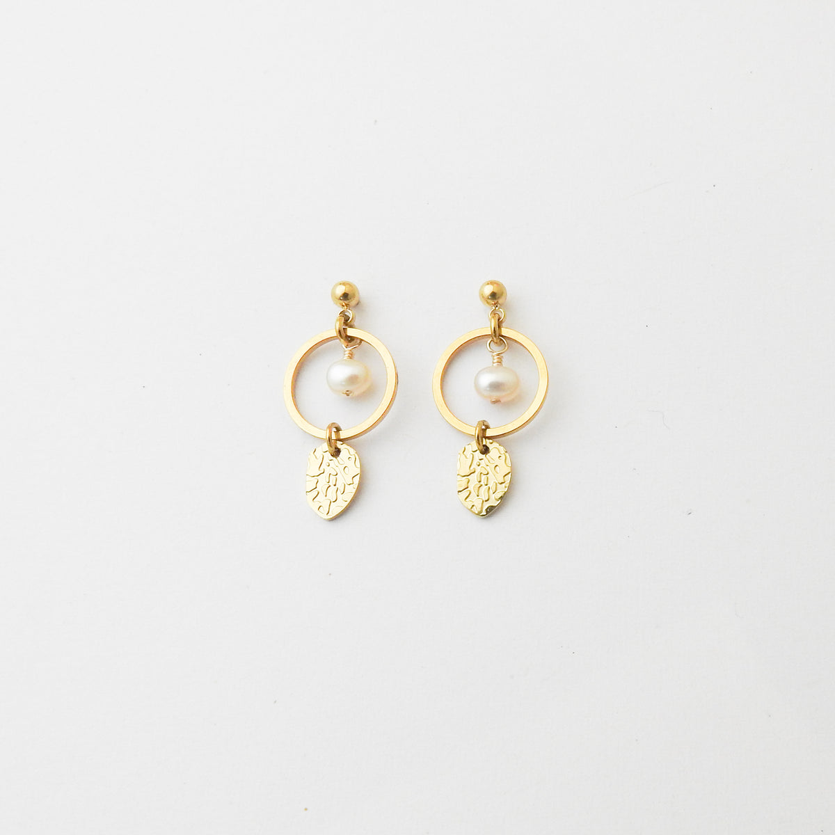 A pair of gold dangle earrings featuring a small white pearl, a gold ring, and a textured leaf-shaped charm, displayed on a clean white background.