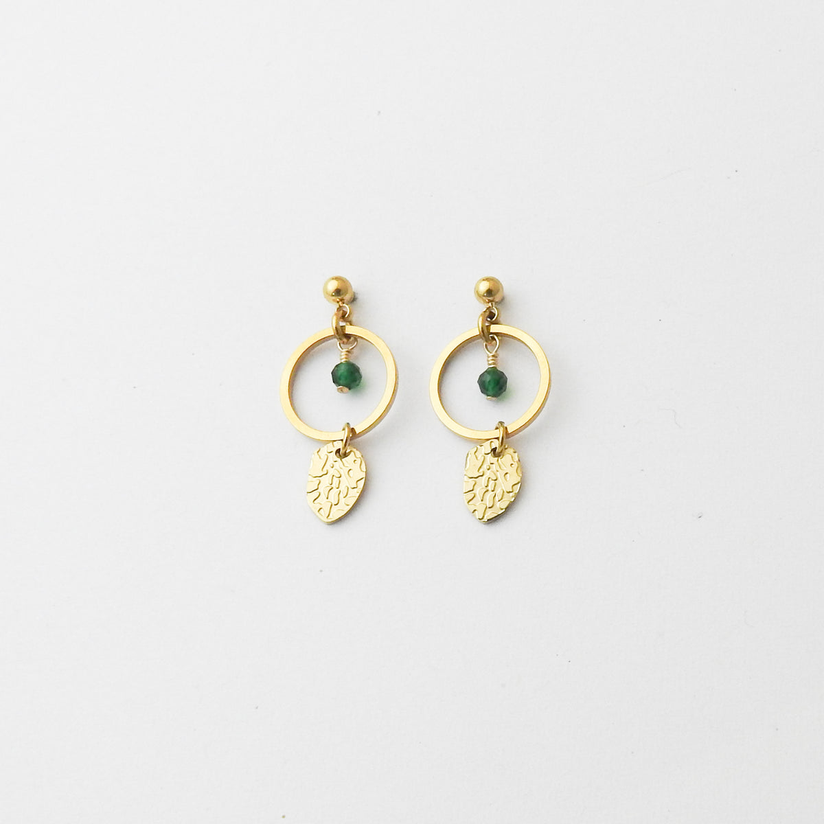 A pair of gold dangle earrings with a small green bead and a textured gold leaf charm, displayed on a clean white background.