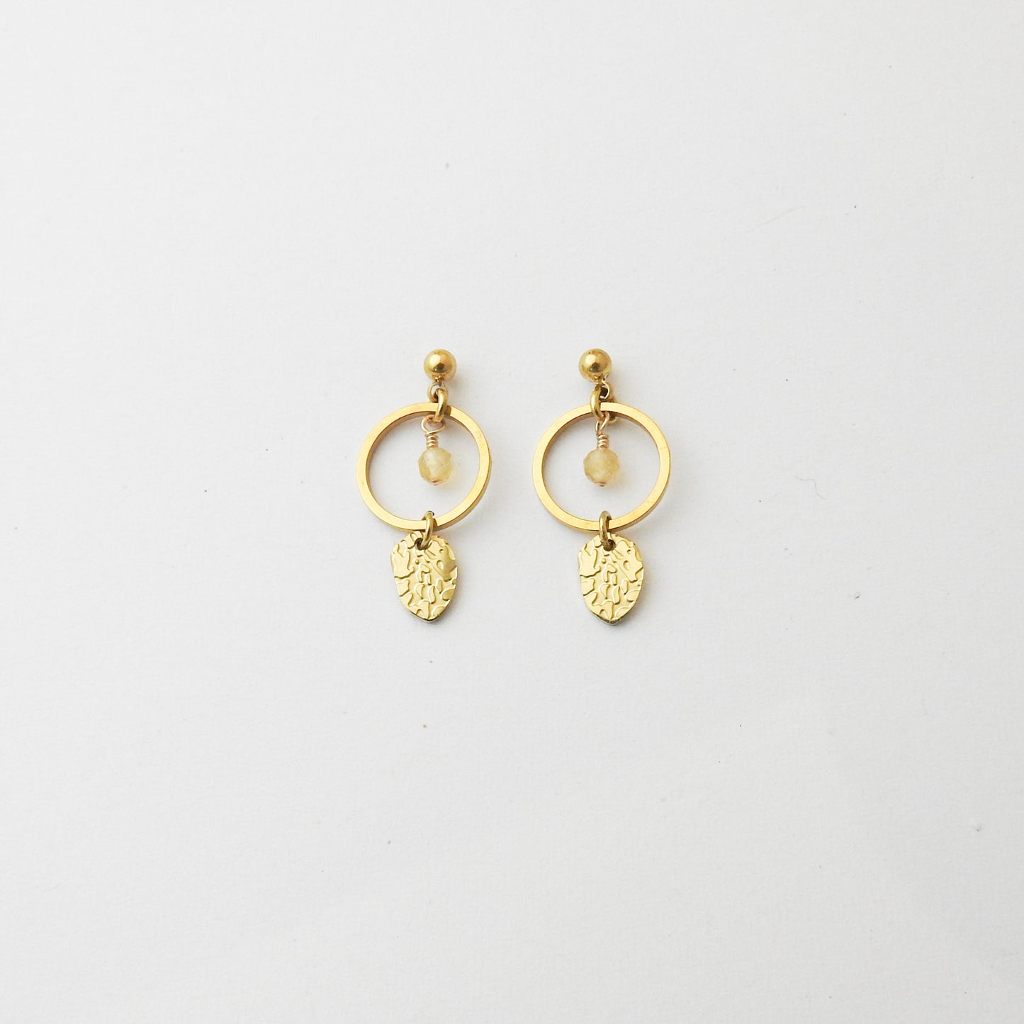 A pair of gold dangle earrings featuring a circular hoop, a small bead, a tiny clear stone, and an engraved leaf-shaped charm, displayed on a clean white background.