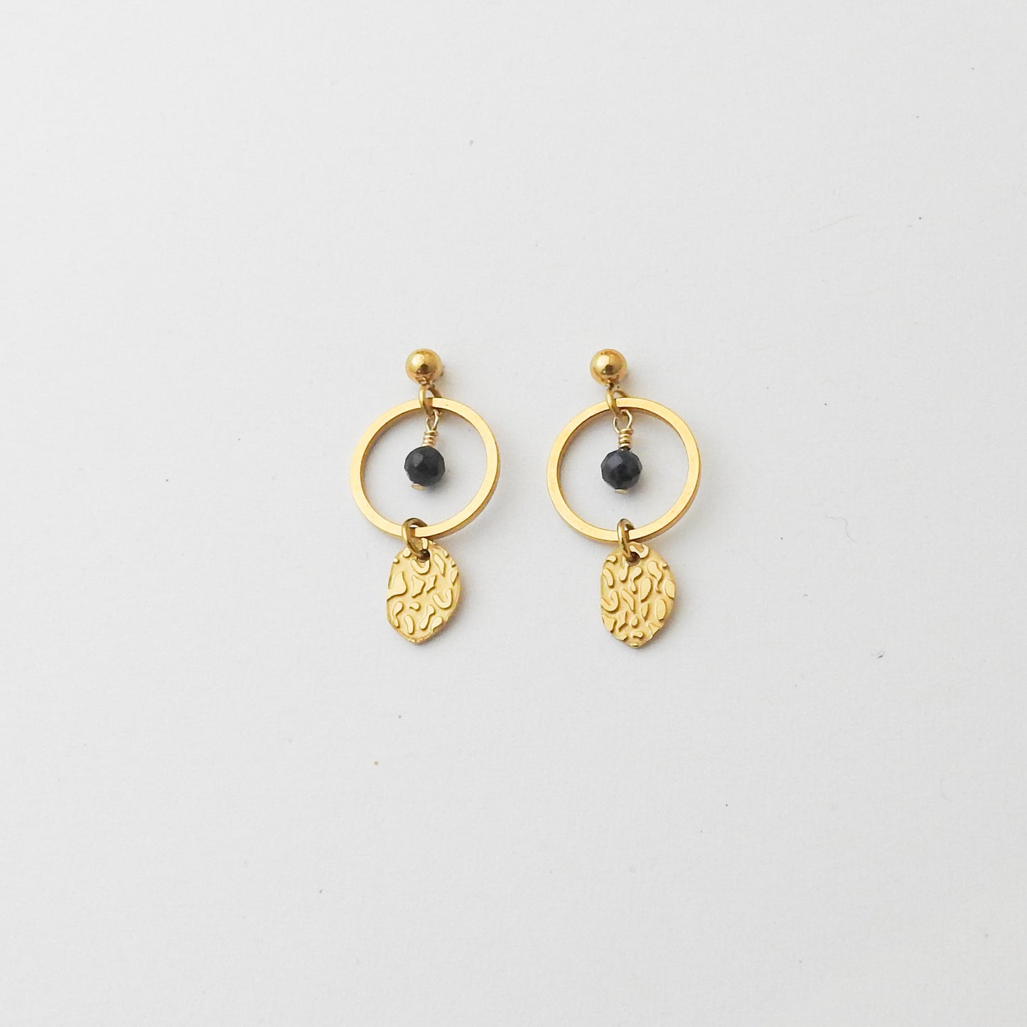 A pair of gold dangle earrings featuring a thin circular hoop, a small black bead, and a textured leaf-shaped charm, displayed on a clean white background.