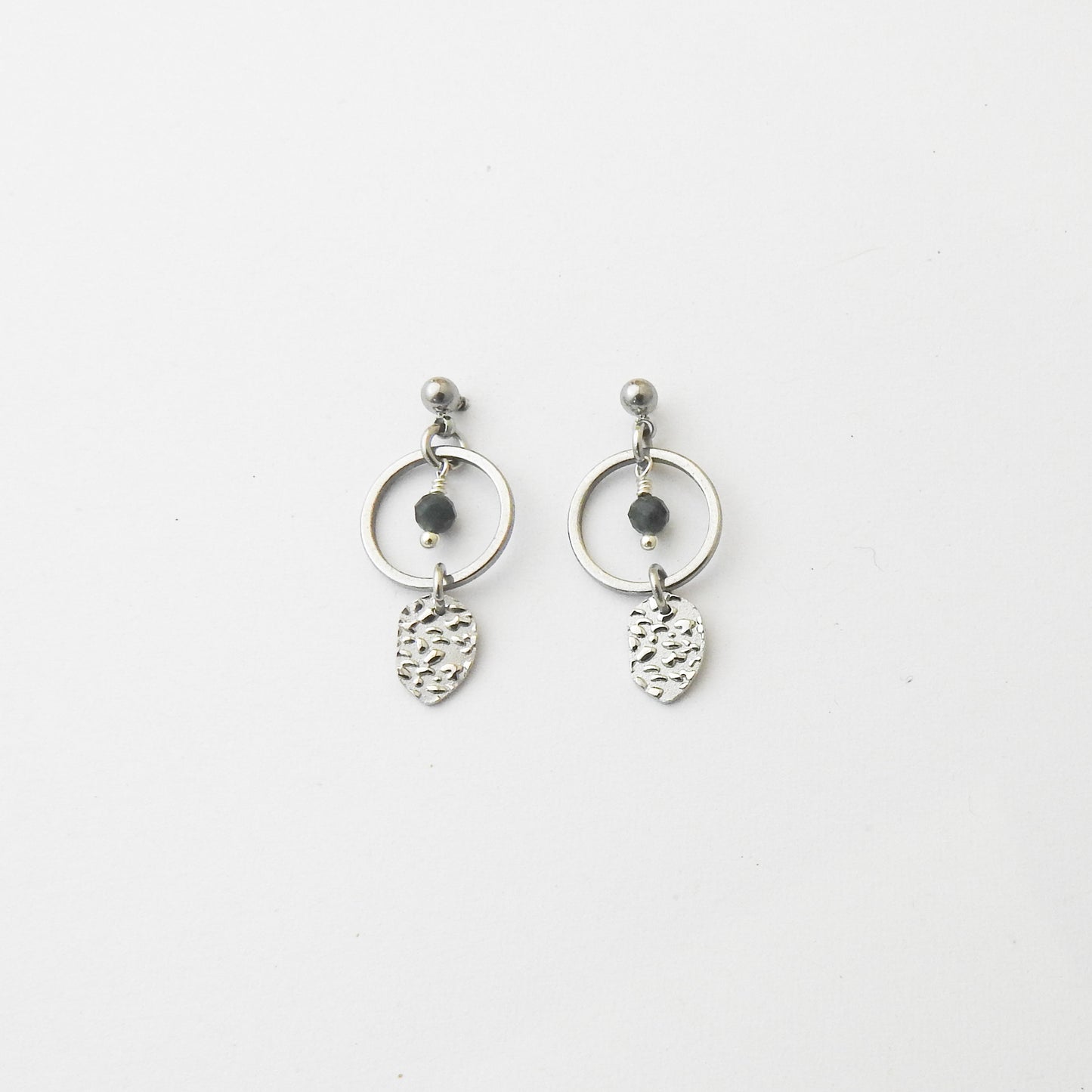 A pair of silver dangle earrings featuring a circular hoop, a small dark bead, and a textured leaf-shaped charm, displayed on a clean white background.