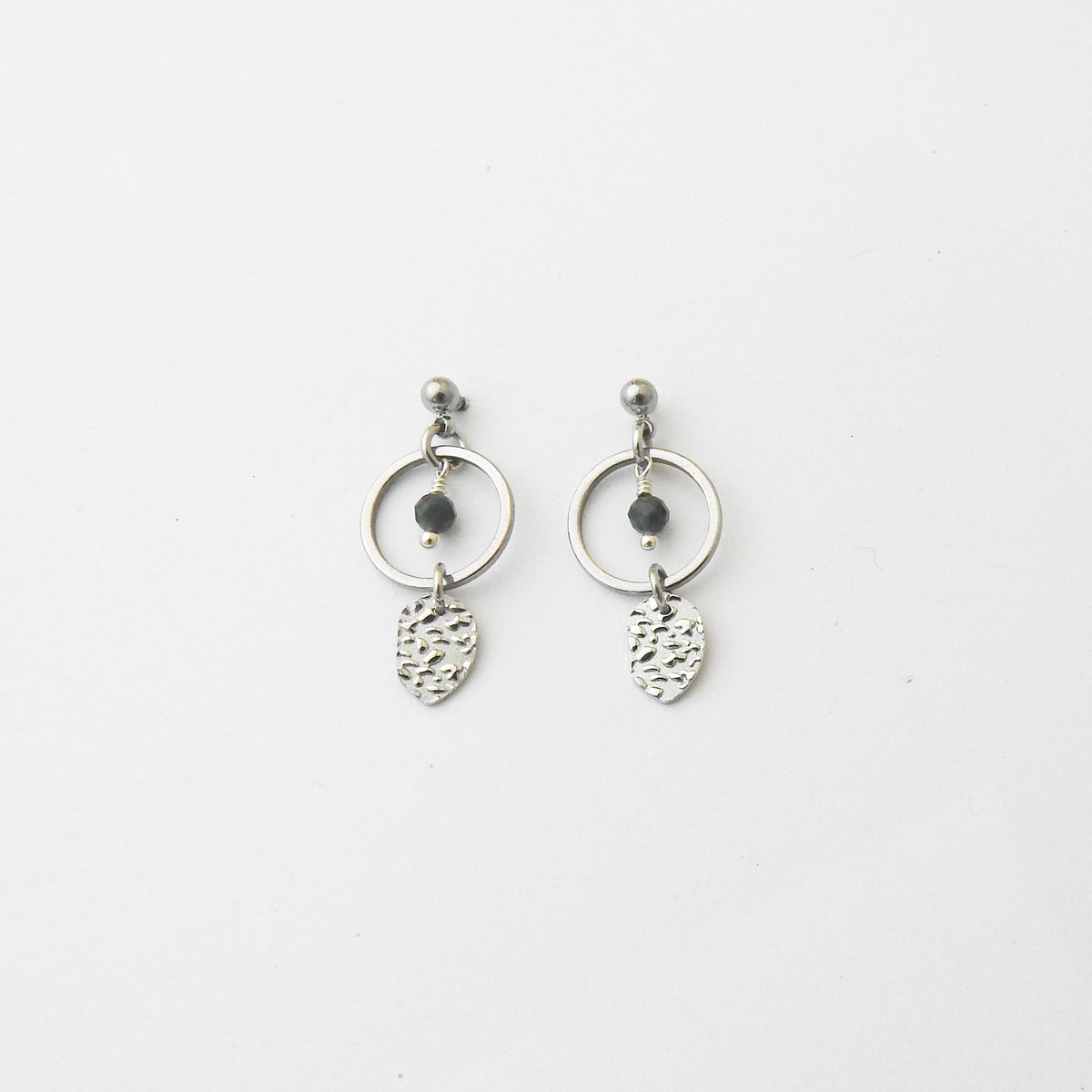 A pair of silver dangle earrings featuring a circular hoop, a small dark bead, and a textured leaf-shaped charm, displayed on a clean white background.