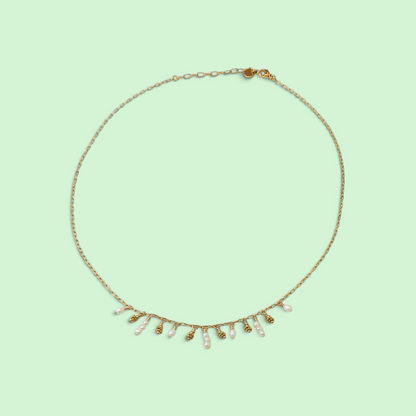 A delicate gold chain necklace with a row of small white pearl-like drops and tiny gold bead accents, displayed on a solid pale green background.