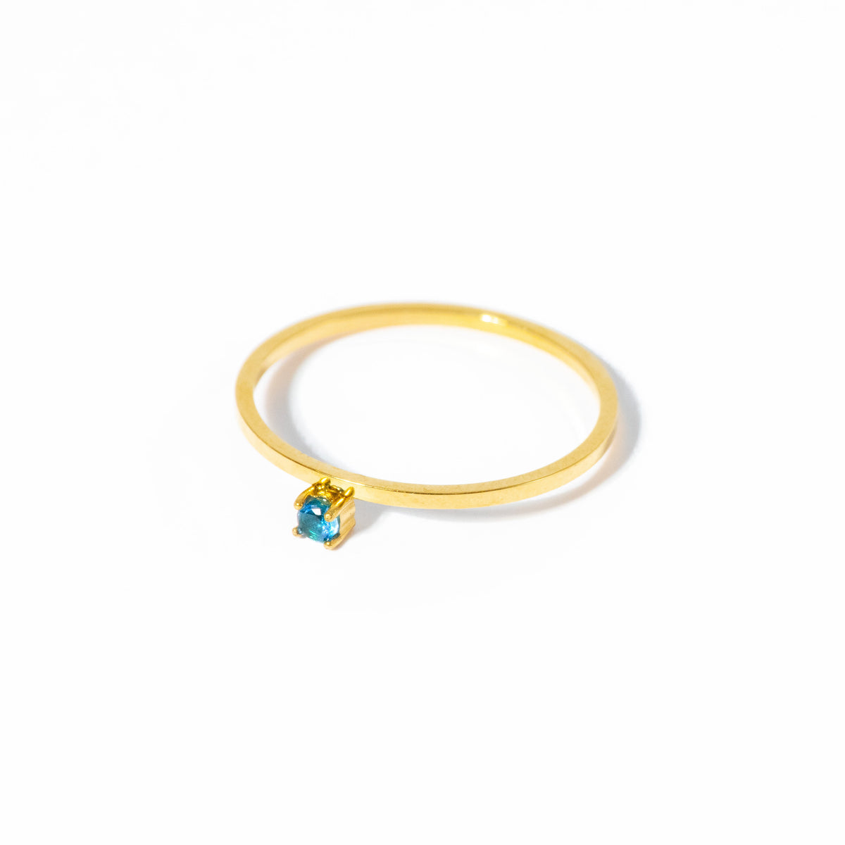 A delicate gold ring featuring a small, bezel-set light blue gemstone, photographed on a clean white background with a soft shadow.