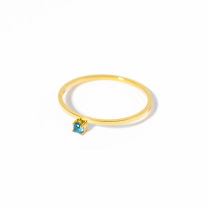 A delicate gold ring featuring a small, bezel-set light blue gemstone, photographed on a clean white background with a soft shadow.