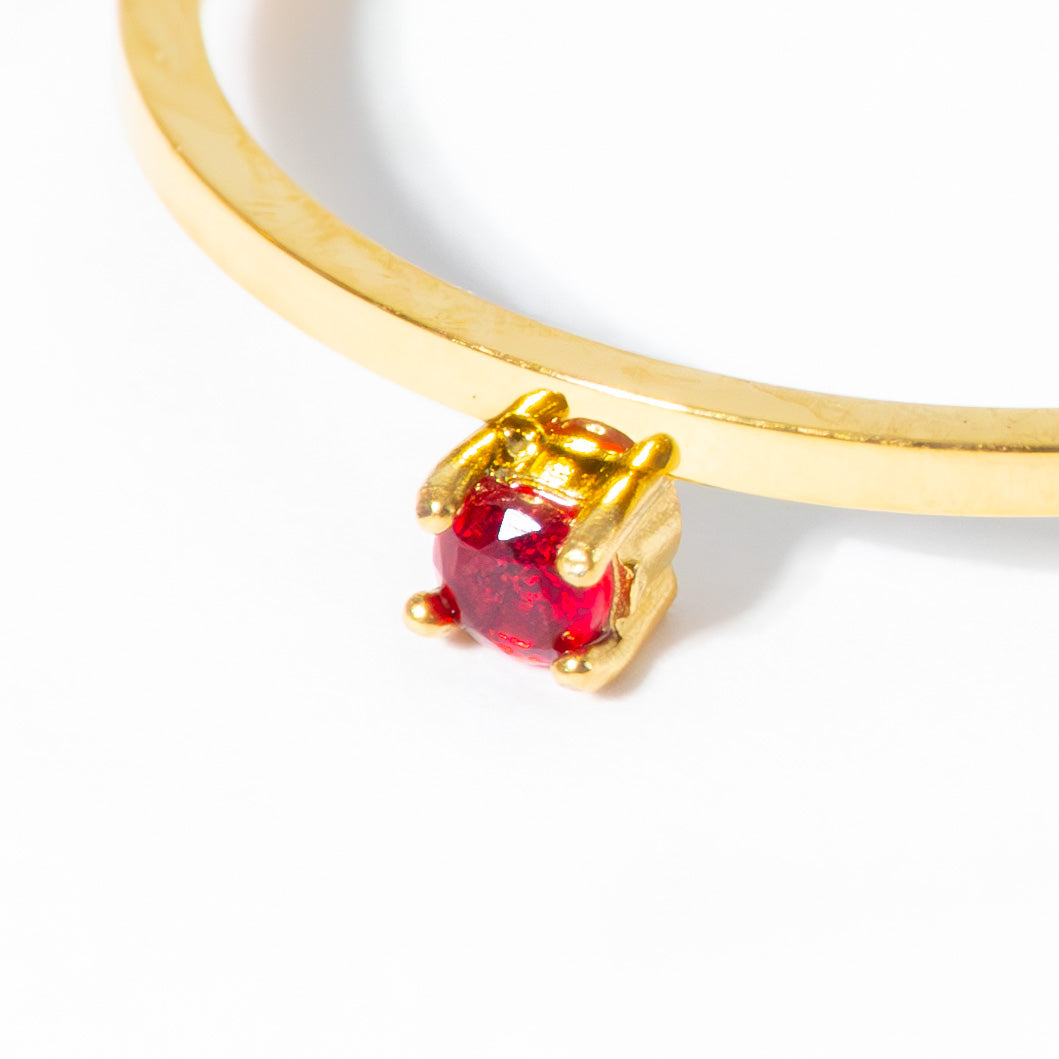 A close-up of a gold ring featuring a small bezel-set round red gemstone on a white background.