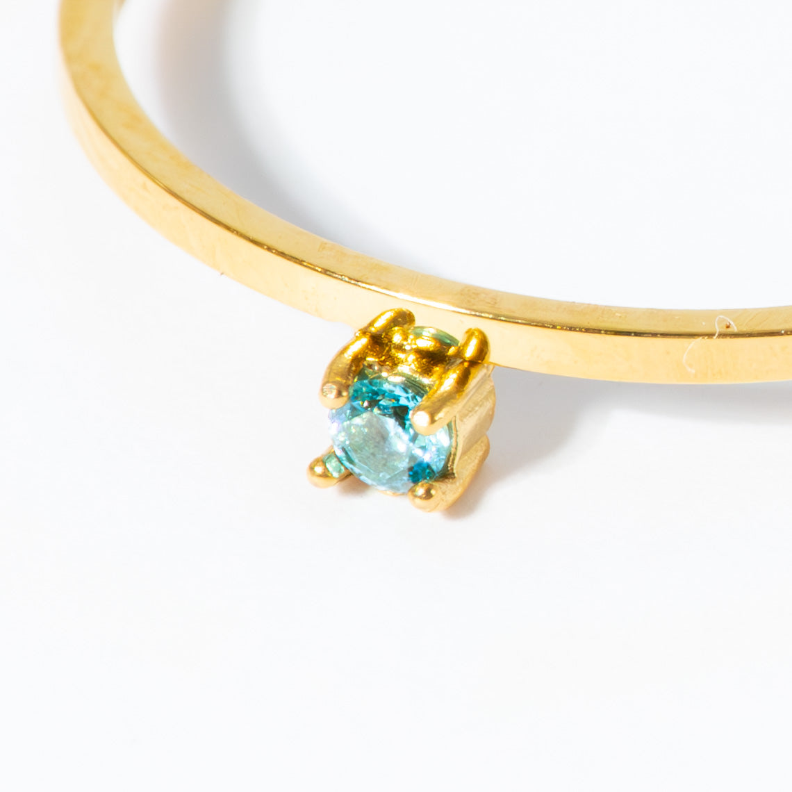 A close-up of a gold bangle bracelet featuring a small gold crown-like setting holding a round light blue gemstone, set against a plain white background.