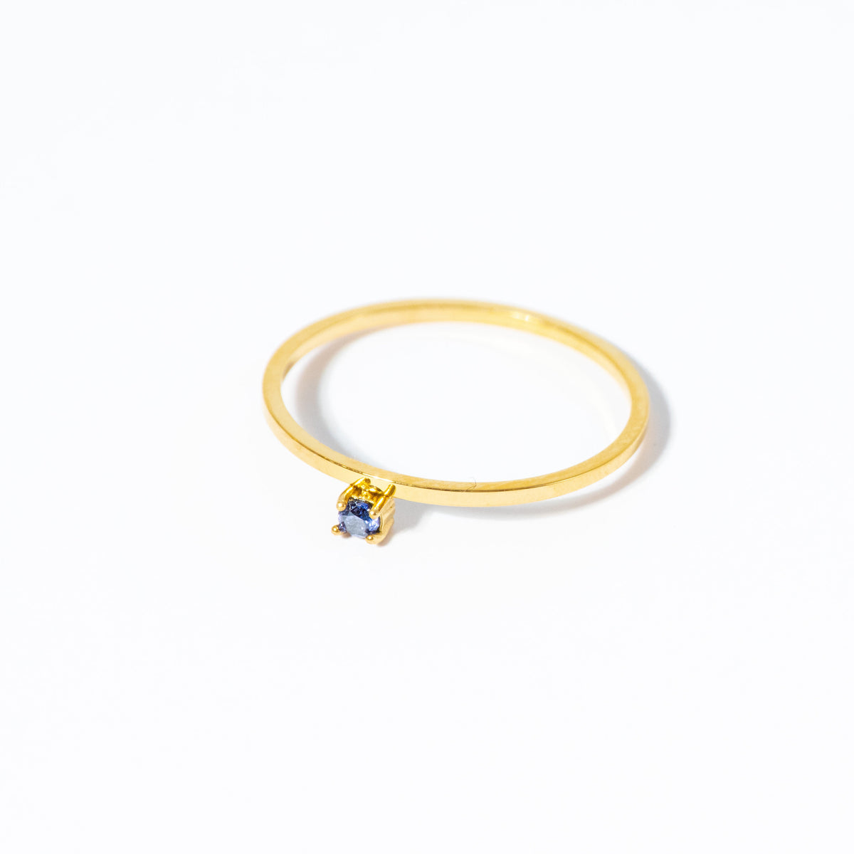 A delicate gold ring featuring a small, round-cut blue gemstone in a prong setting, photographed on a clean white background.