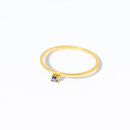 A delicate gold ring featuring a small, round-cut blue gemstone in a prong setting, photographed on a clean white background.