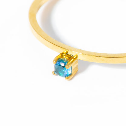 A close-up of a gold bangle bracelet featuring a small, square-cut blue gemstone in a gold prong setting, shot against a clean white background.