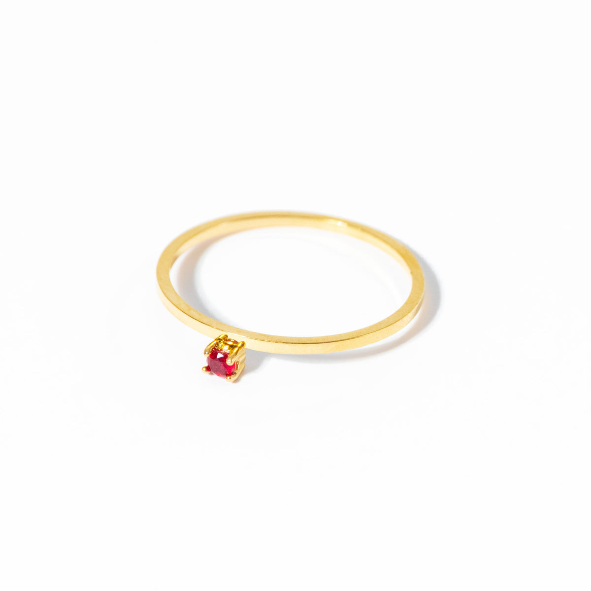 A delicate gold ring featuring a small, round-cut red gemstone in a prong setting, photographed on a clean white background.