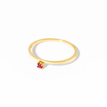 A delicate gold ring featuring a small, round-cut red gemstone in a prong setting, photographed on a clean white background.
