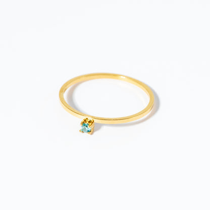 A delicate gold ring featuring a small, bezel-set light blue heart-shaped gemstone, photographed on a clean white background.