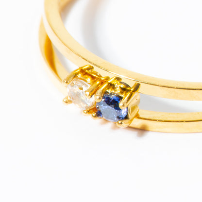 A close-up of a gold double-band ring featuring a triple gemstone cluster with a clear round stone, a blue oval sapphire, and a smaller clear round stone in a gold prong setting, on a white background.