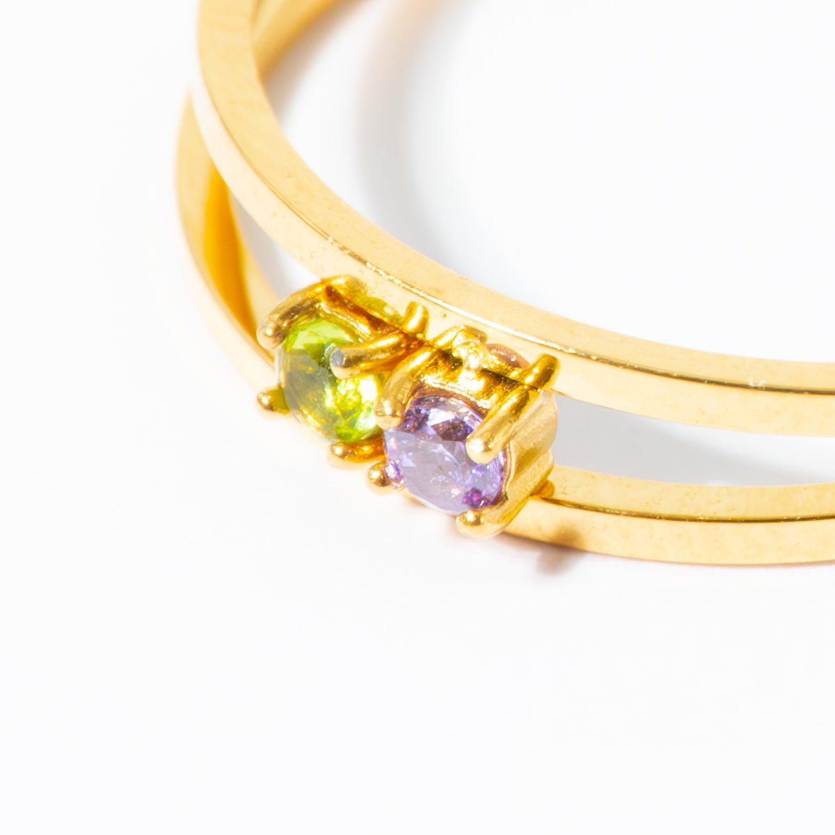A close-up of a gold double-band ring featuring two round gemstones, one green and one purple, set against a plain white background.
