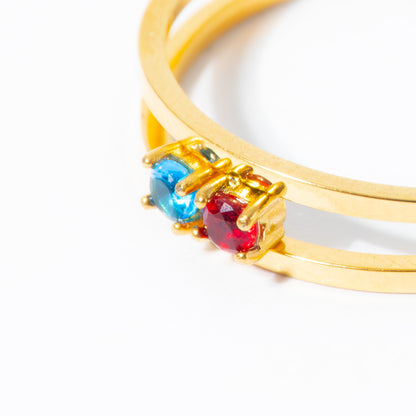 A close-up of a gold double-band ring featuring two round gemstones, one blue and one red, set in a prong-style clasp on a white background.