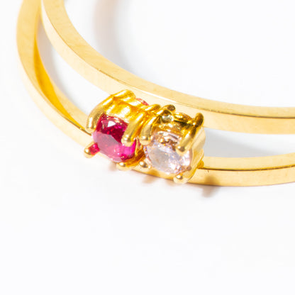 A close-up of a gold double-band ring featuring a pink gemstone and a clear round-cut gemstone in a prong setting, on a white background.