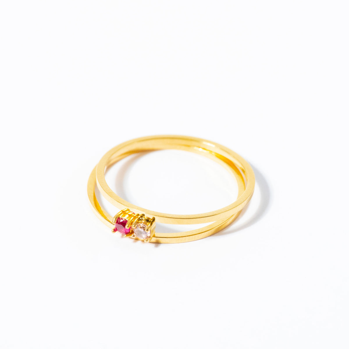 A gold double-band ring with a small heart-shaped cluster of pink and clear gemstones, photographed on a clean white background.