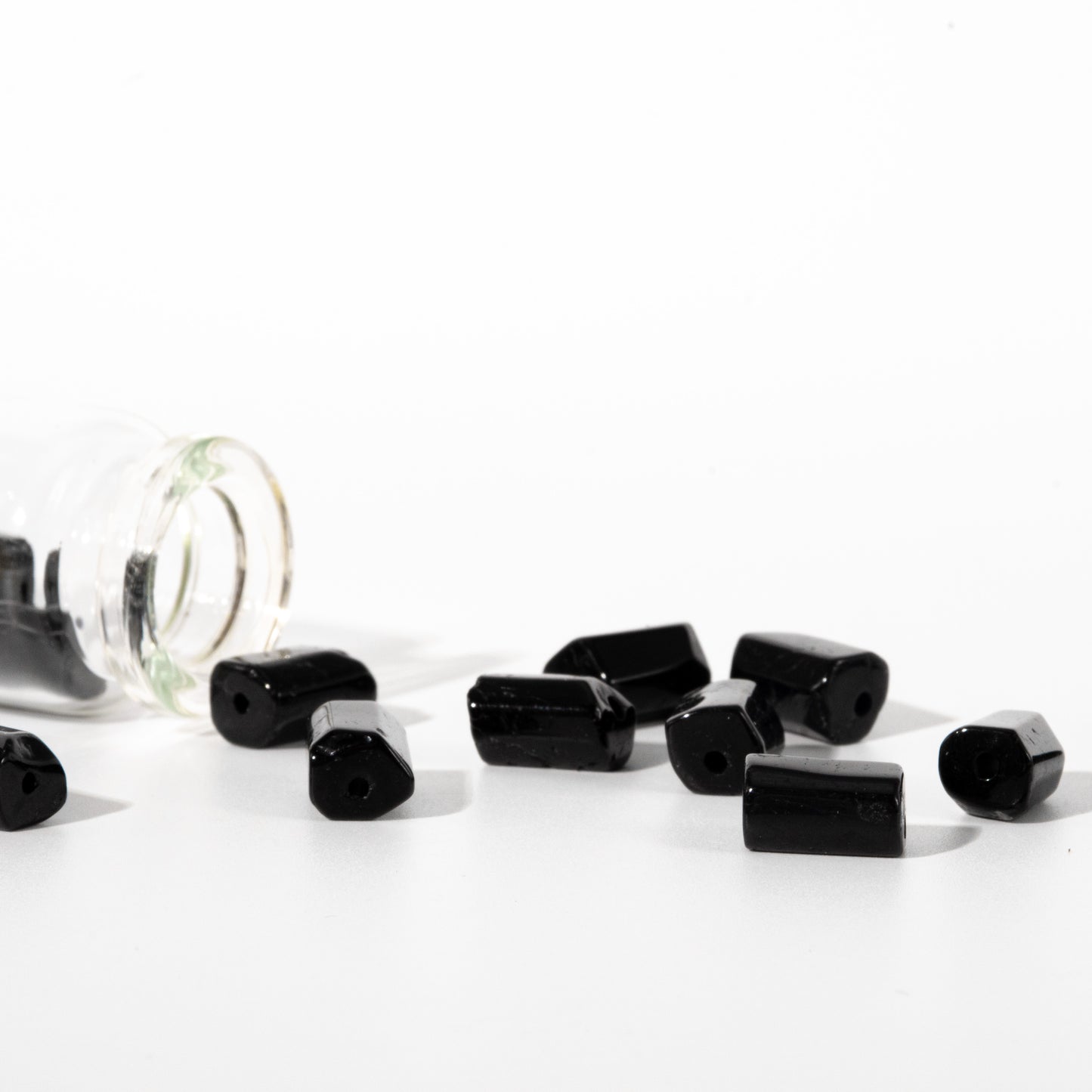 Several glossy black cylindrical beads with flat ends scattered on a white surface, with a clear glass jar tipped over on the left.