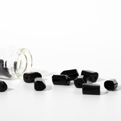 Several glossy black cylindrical beads with flat ends scattered on a white surface, with a clear glass jar tipped over on the left.