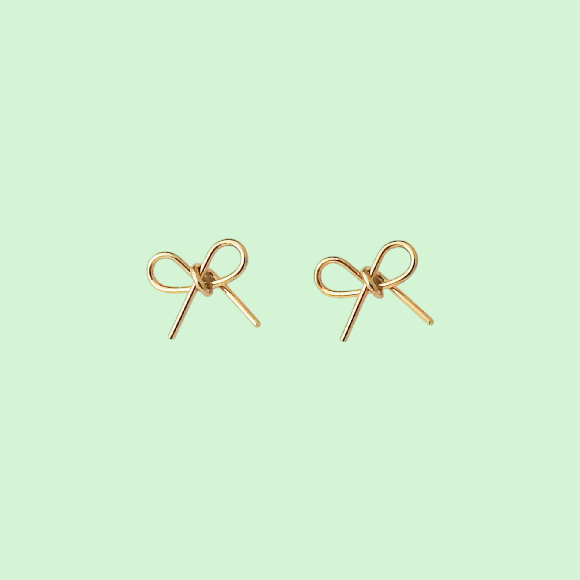 A pair of small gold bow-shaped stud earrings on a solid pale mint green background.