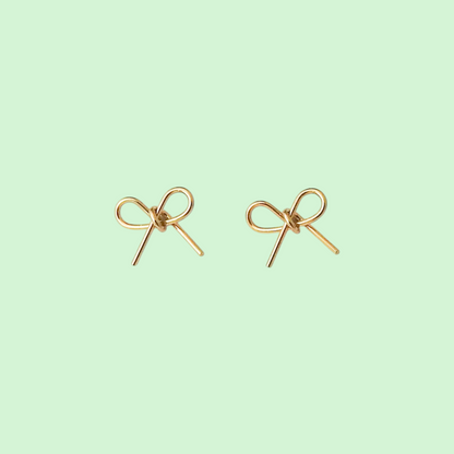 A pair of small gold bow-shaped stud earrings on a solid pale mint green background.