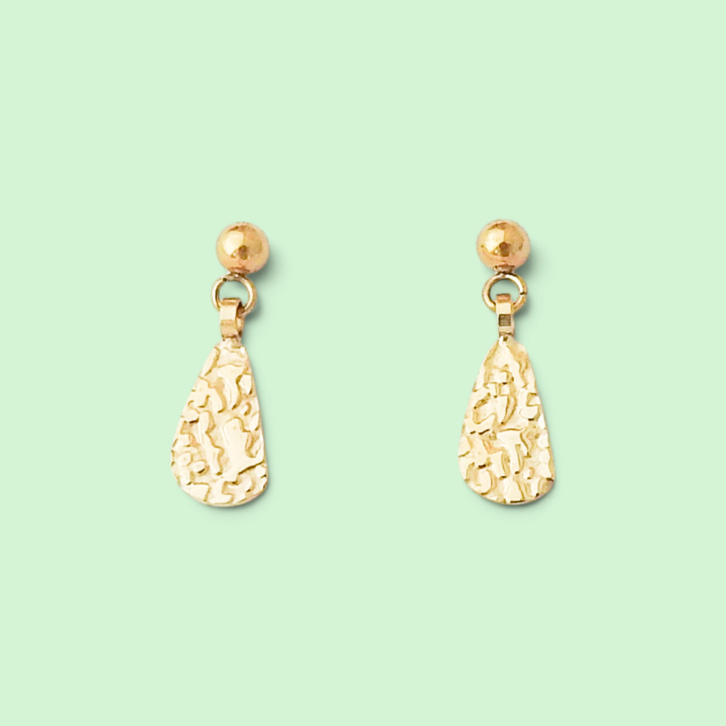 A pair of gold dangle earrings with textured teardrop-shaped drops and round stud tops, displayed on a solid pale mint green background.