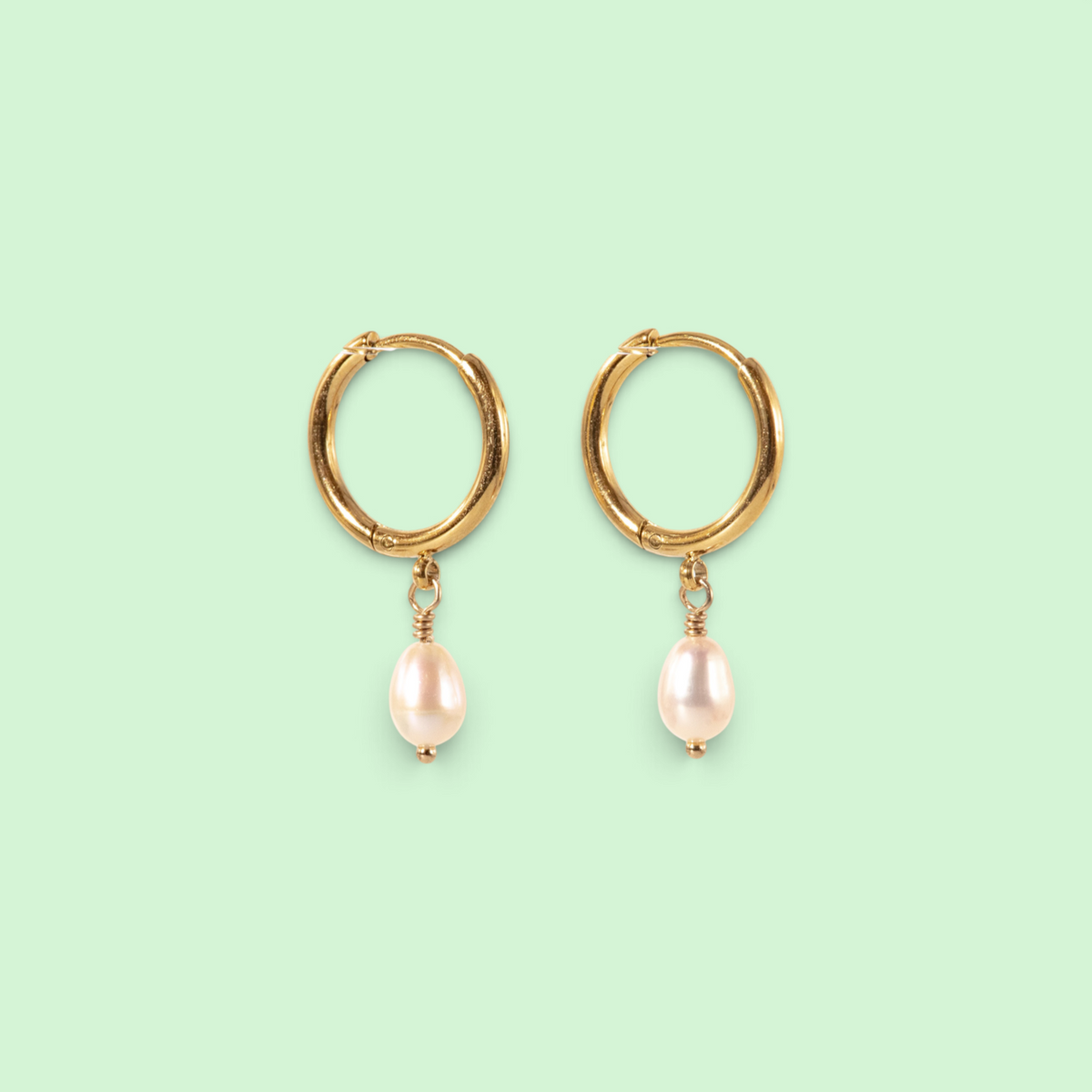 A pair of gold huggie hoop earrings with dangling baroque pearl charms, shown on a solid pale mint green background.