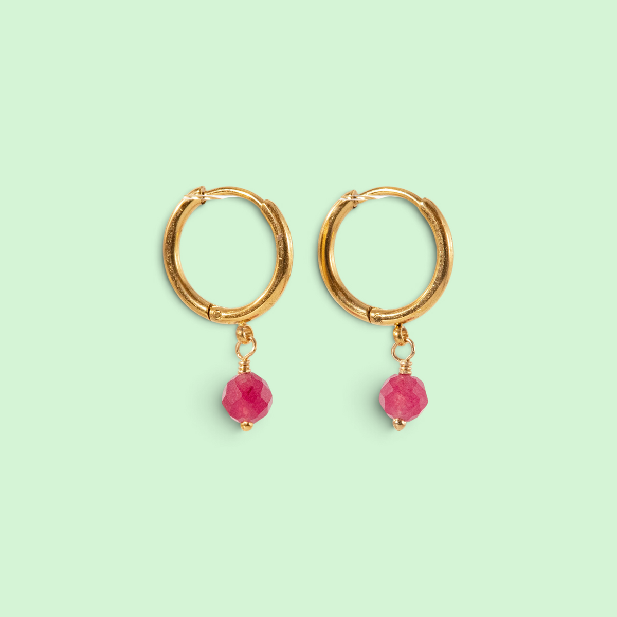 A pair of gold hoop earrings with faceted pink gemstone dangles, displayed on a solid pale mint green background.