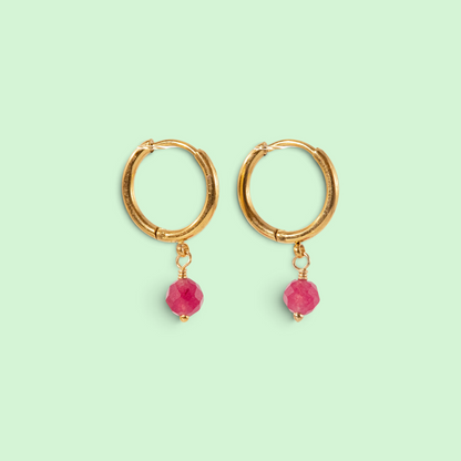 A pair of gold hoop earrings with faceted pink gemstone dangles, displayed on a solid pale mint green background.