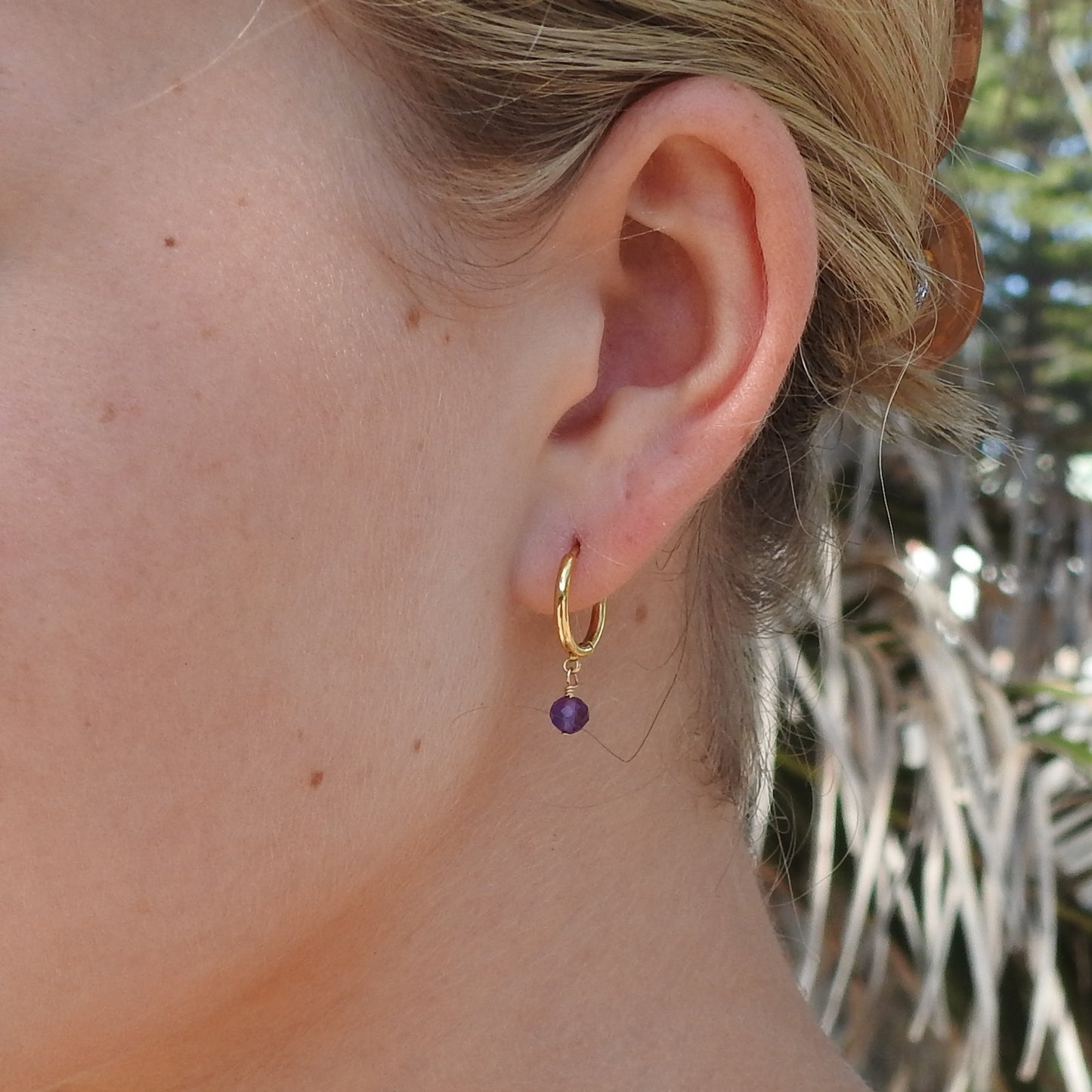 A close-up of a person's ear wearing a small gold hoop earring with a dangling purple bead, set against an outdoor background with blurred foliage.