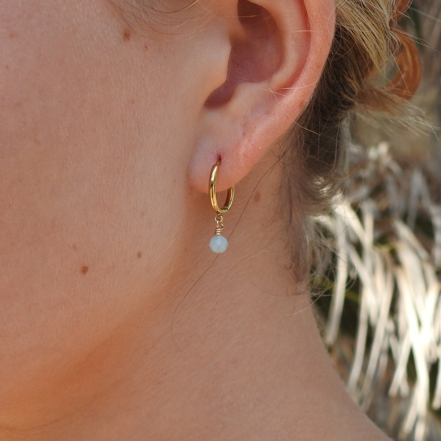 A close-up of a person's ear wearing a small gold hoop earring with a dangling light blue pearl bead.
