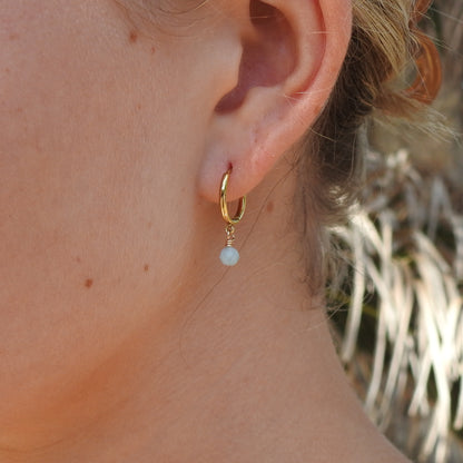 A close-up of a person's ear wearing a small gold hoop earring with a dangling light blue pearl bead.