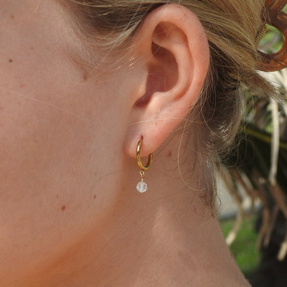 A close-up of a person's ear wearing a small gold hoop earring with a dangling clear faceted bead.