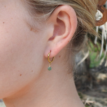 A close-up of a person's ear wearing a small gold hoop earring with a dangling green bead charm, against a softly blurred outdoor background.