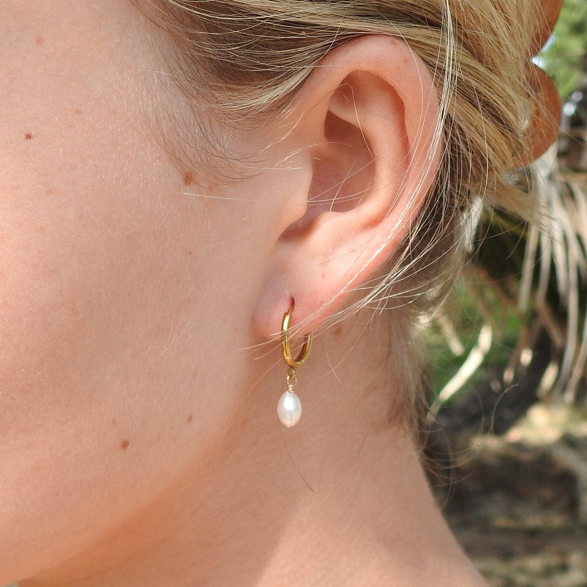 A close-up of a person's ear wearing a small gold hoop earring with a dangling white pearl, photographed outdoors with a blurred natural background.